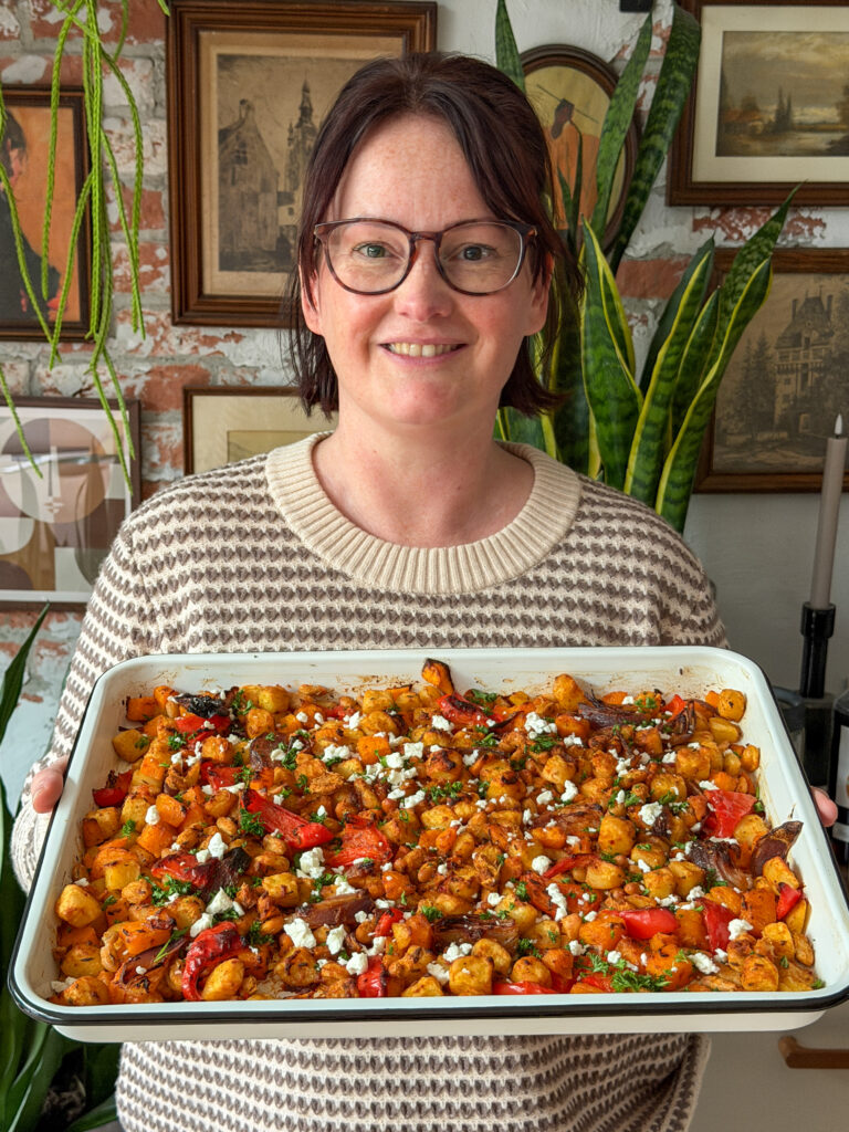 Marie Louise houdt een ovenschaal vast met zelfgemaakte traybake van pompoen, krieltjes, harissa en feta
